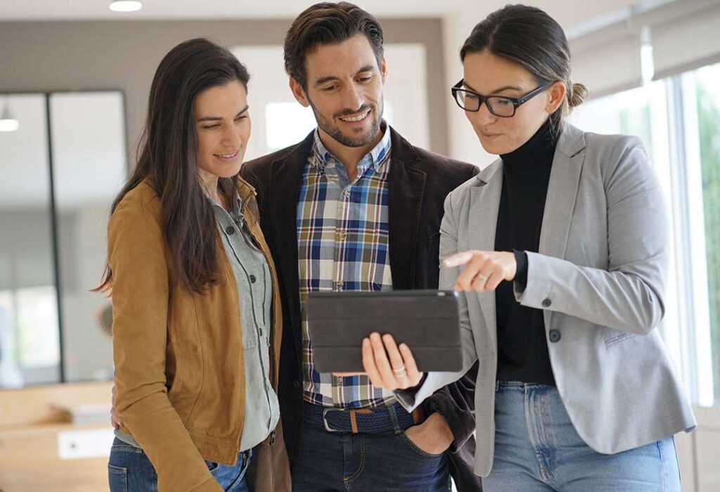 A young couple discusses paperwork with a real estate agent in a well-lit, residential living room.