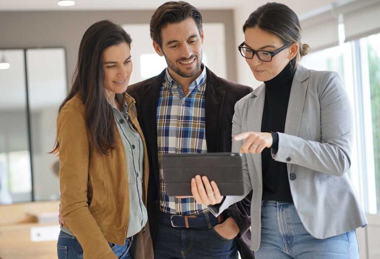 A young couple discusses paperwork with a real estate agent in a well-lit, residential living room.